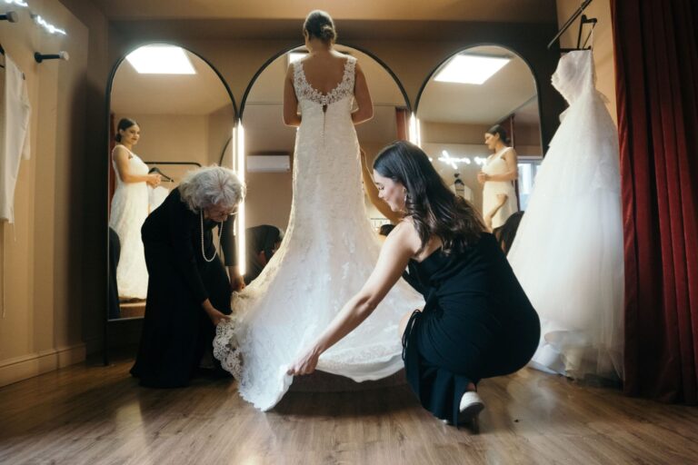 Due donne sistemano lo strascico in pizzo di una sposa durante la prova dell'abito, davanti agli specchi